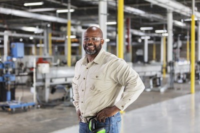 A mature African-American man working in a plastics factory. He is standing on the factory floor with his hands on his hips, smiling at the camera.