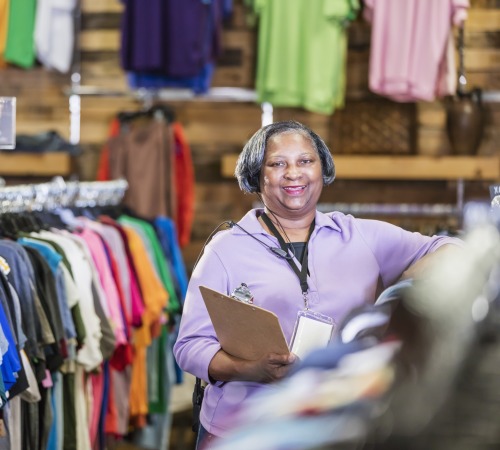 A senior African-American woman in her 60s working in a clothing store. She is standing in the menswear department at a rack of clothing, holding a clipboard.